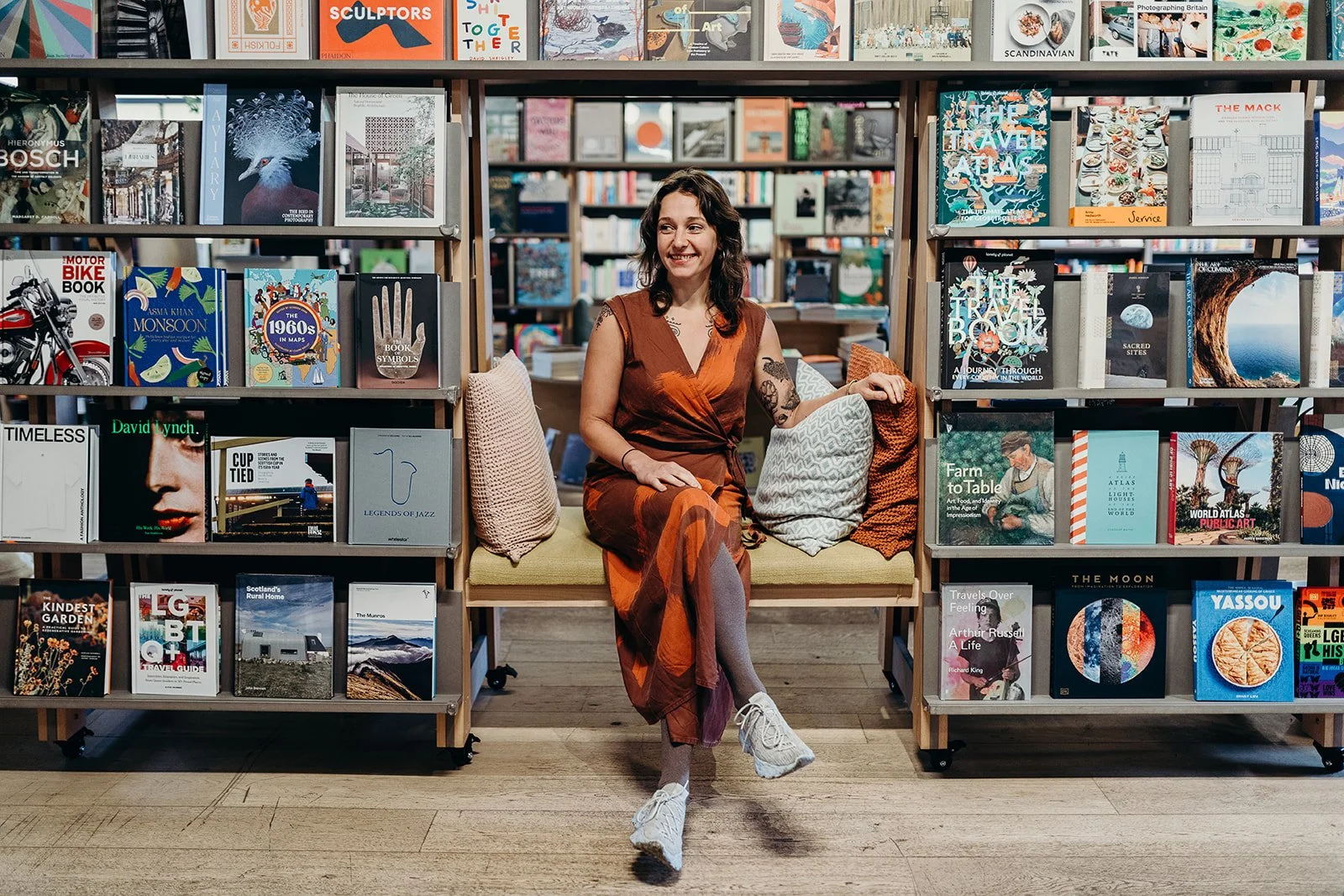 Lily Asch sitting on a bench between two bookshelves filled with books in a bookstore. The woman is smiling, wearing an orange and brown dress, and has dark wavy hair. She is sitting with her legs crossed, with decorative pillows beside her.