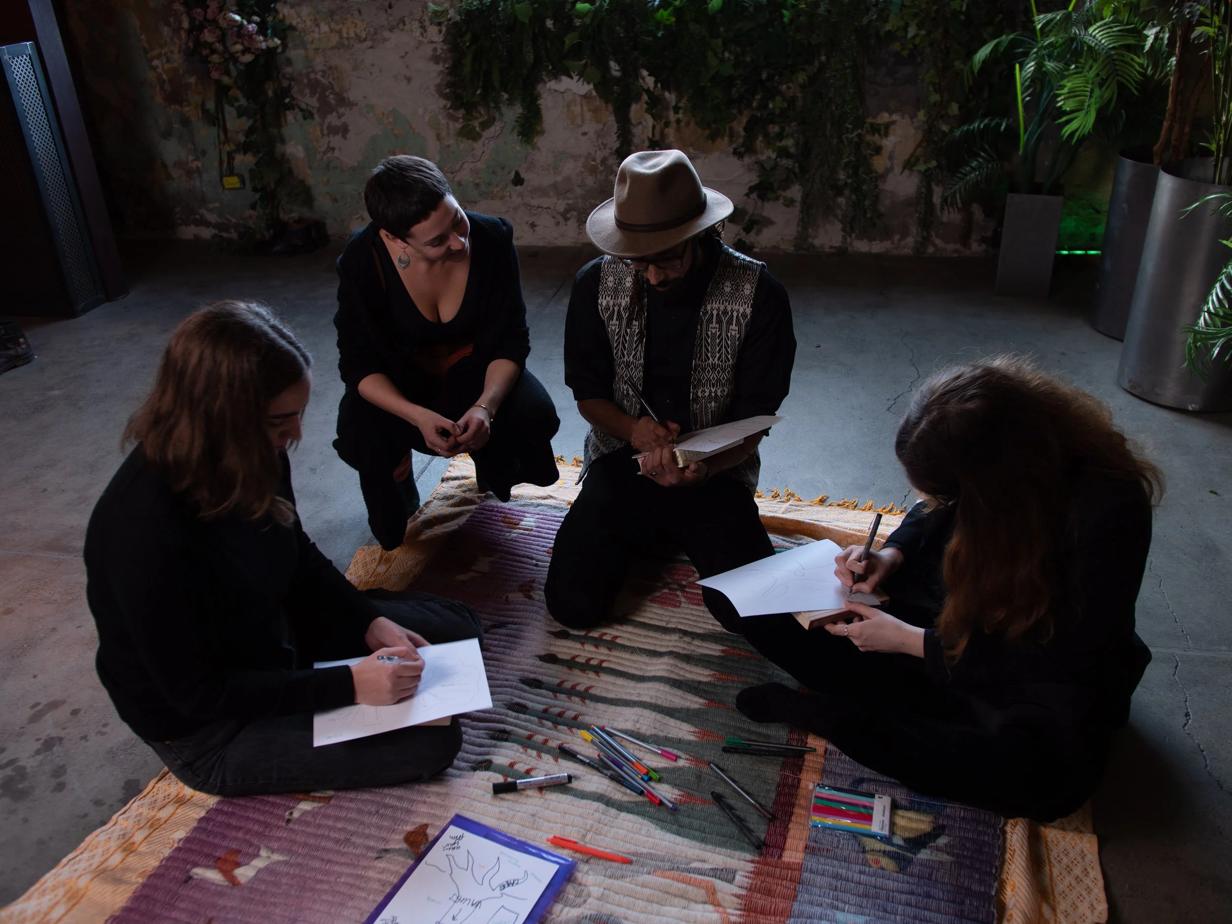 Five people sitting on a colorful woven rug making sketches and notes with various art supplies, in a room with potted plants and a textured wall.