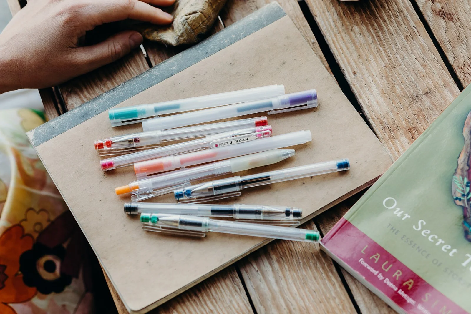 A collection of colorful pens and markers laid out on a beige notebook on a wooden table, with a book titled 'Our Secret' nearby.