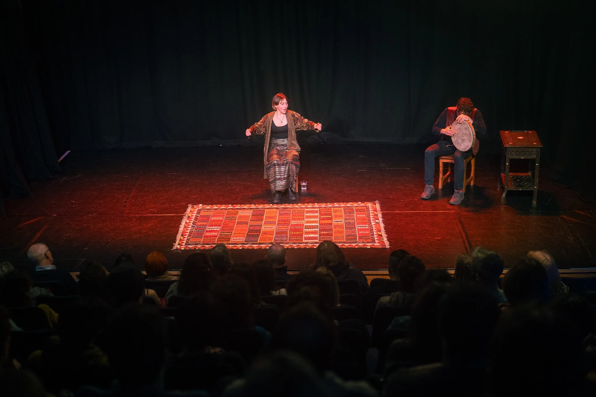 Two performers on stage, one woman sitting and speaking, one man sitting and playing a drum, audience watching in a theater with dark curtains.