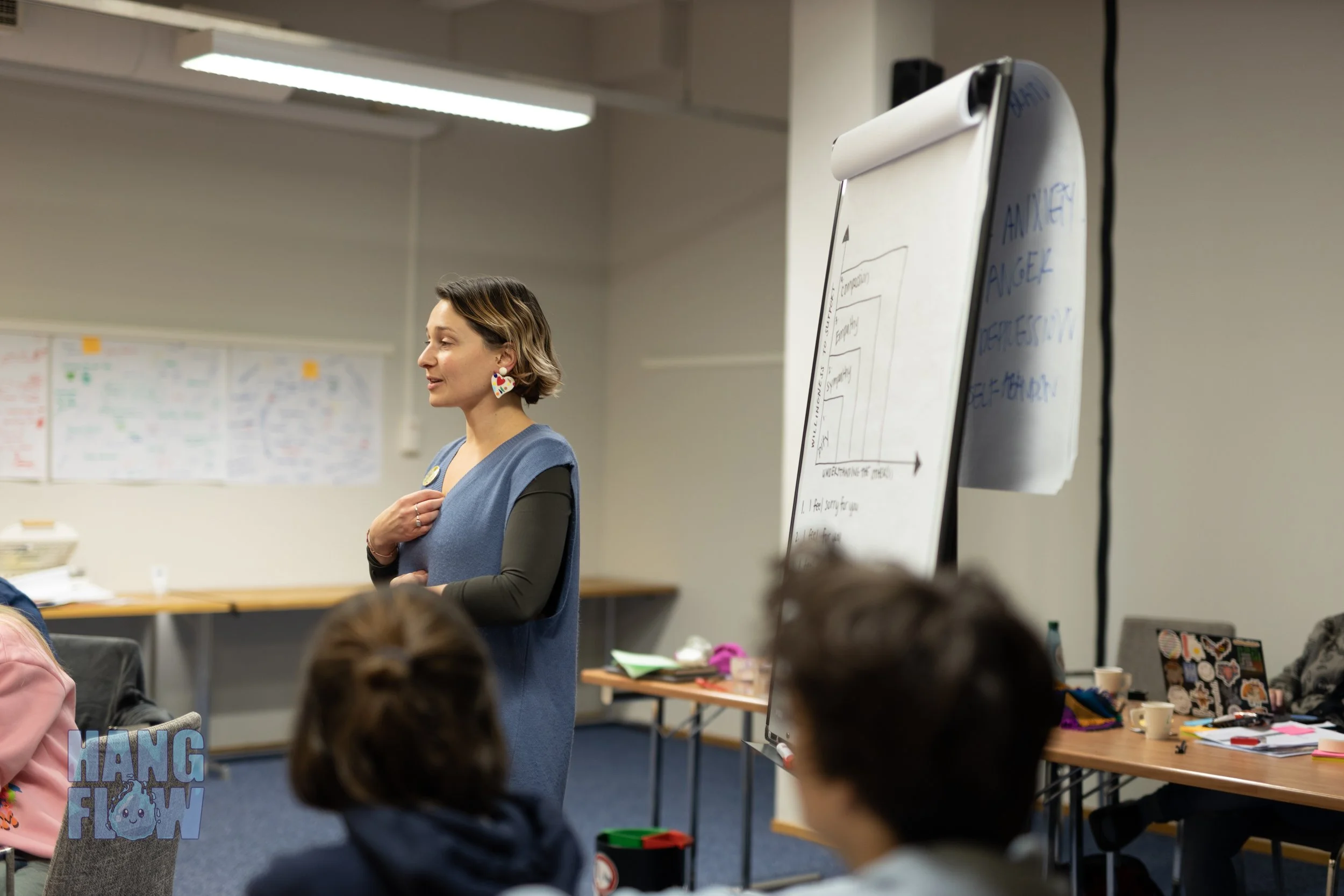 Lily Asch in a blue vest and colorful earrings is speaking to an audience in a conference room, with a whiteboard and tables with papers and cups in the background.