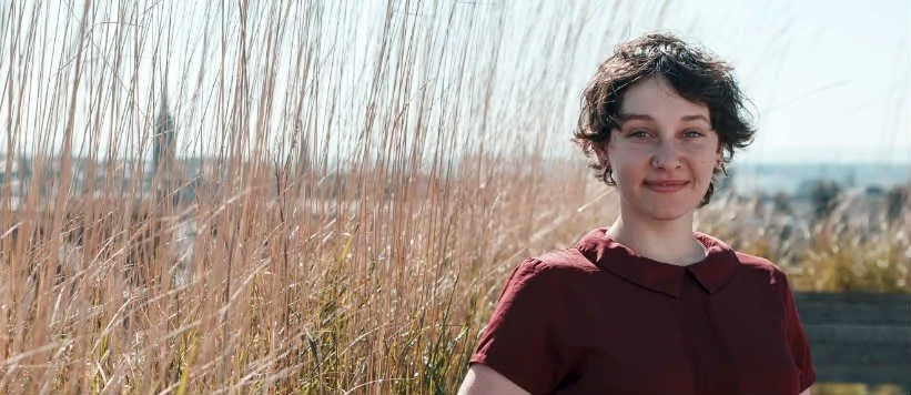 A young woman with short, curly hair and a maroon shirt standing outdoors near tall grasses, with a clear sky and distant cityscape in the background.