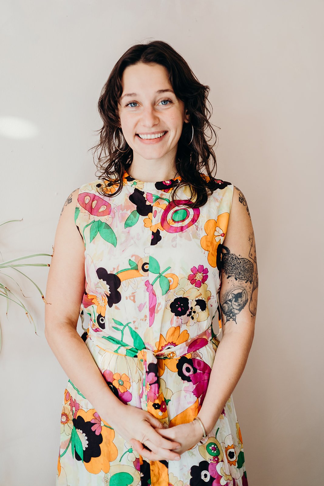 Lily Asch with curly dark hair, wearing a colorful floral sleeveless dress, standing against a plain wall, smiling at the camera.