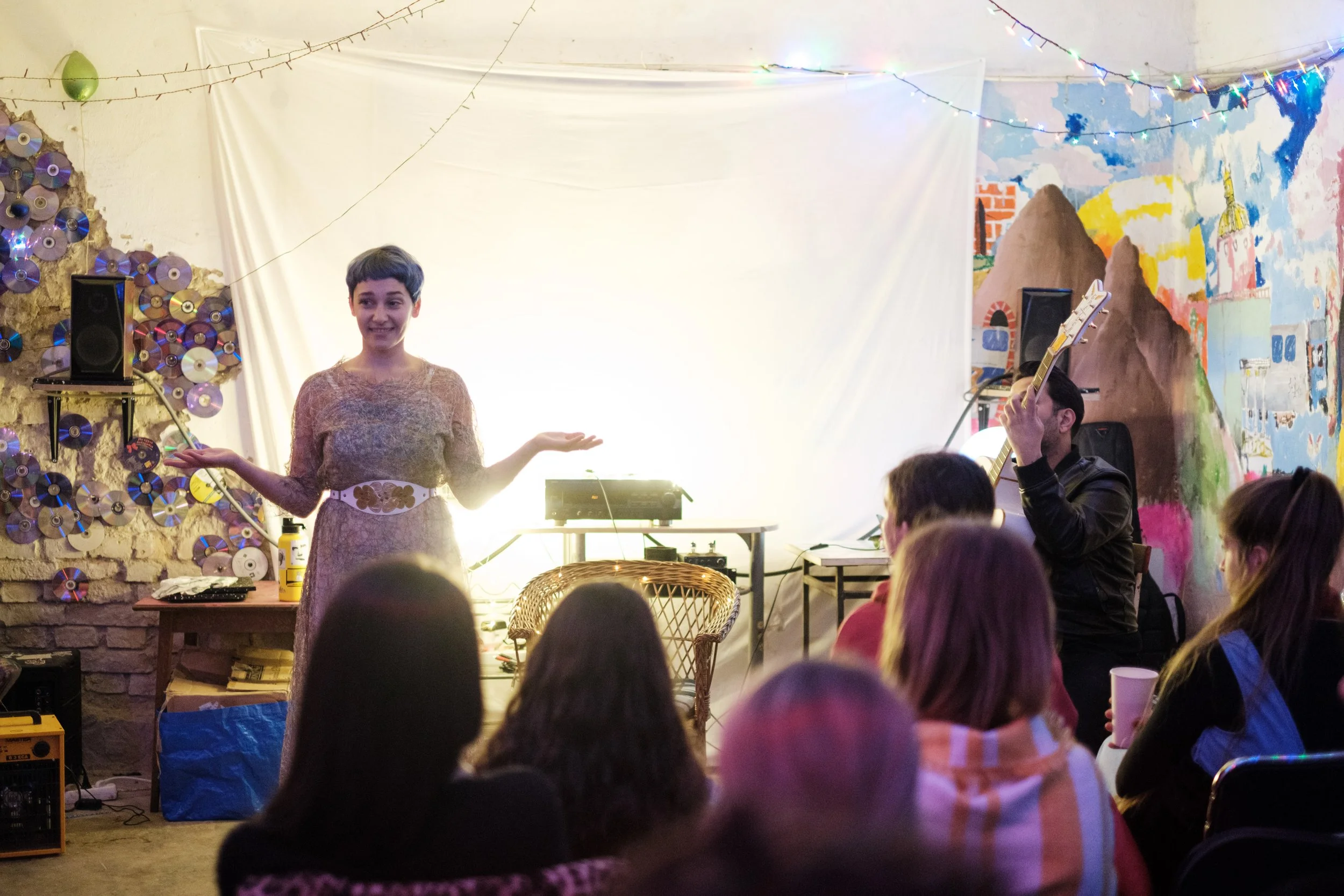 A woman with short hair and a patterned dress stands in front of a small audience with her arms raised, smiling, in a decorated indoor space with string lights and a colorful mural. A man playing a guitar is seated to the right of the woman.