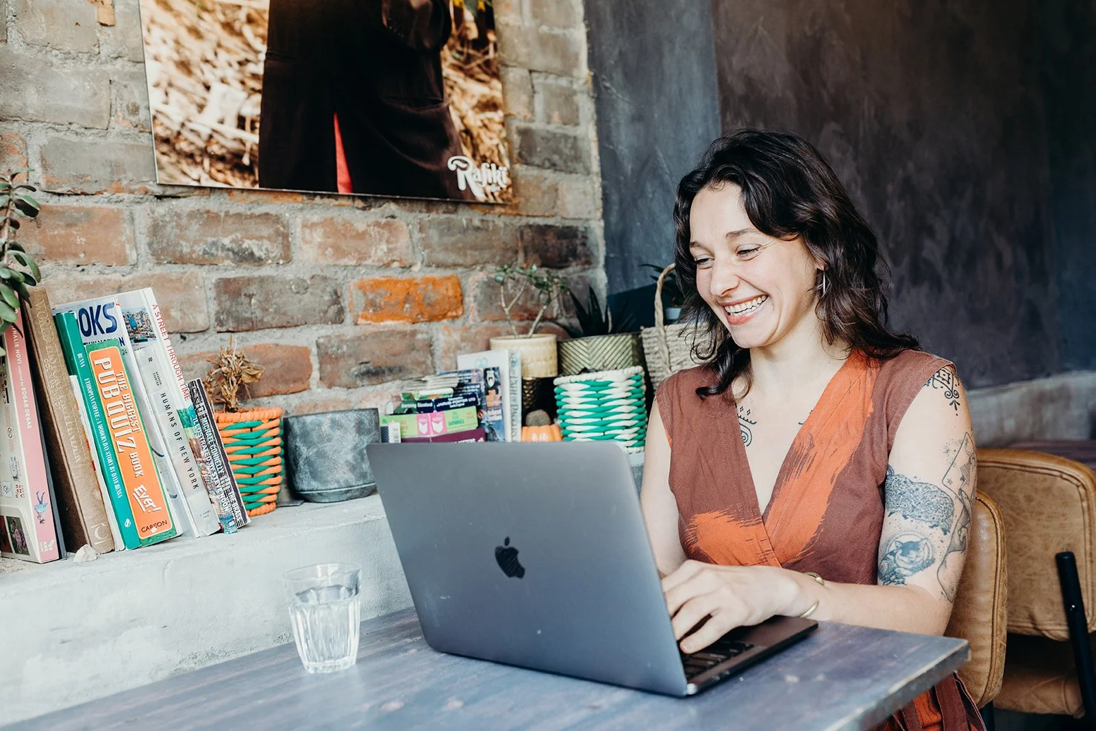 A woman with dark wavy hair, tattoos on her arms, and wearing a brown sleeveless top, smiling while sitting at a table working on a laptop in a cozy cafe with a brick wall and shelves filled with books and plants in the background.