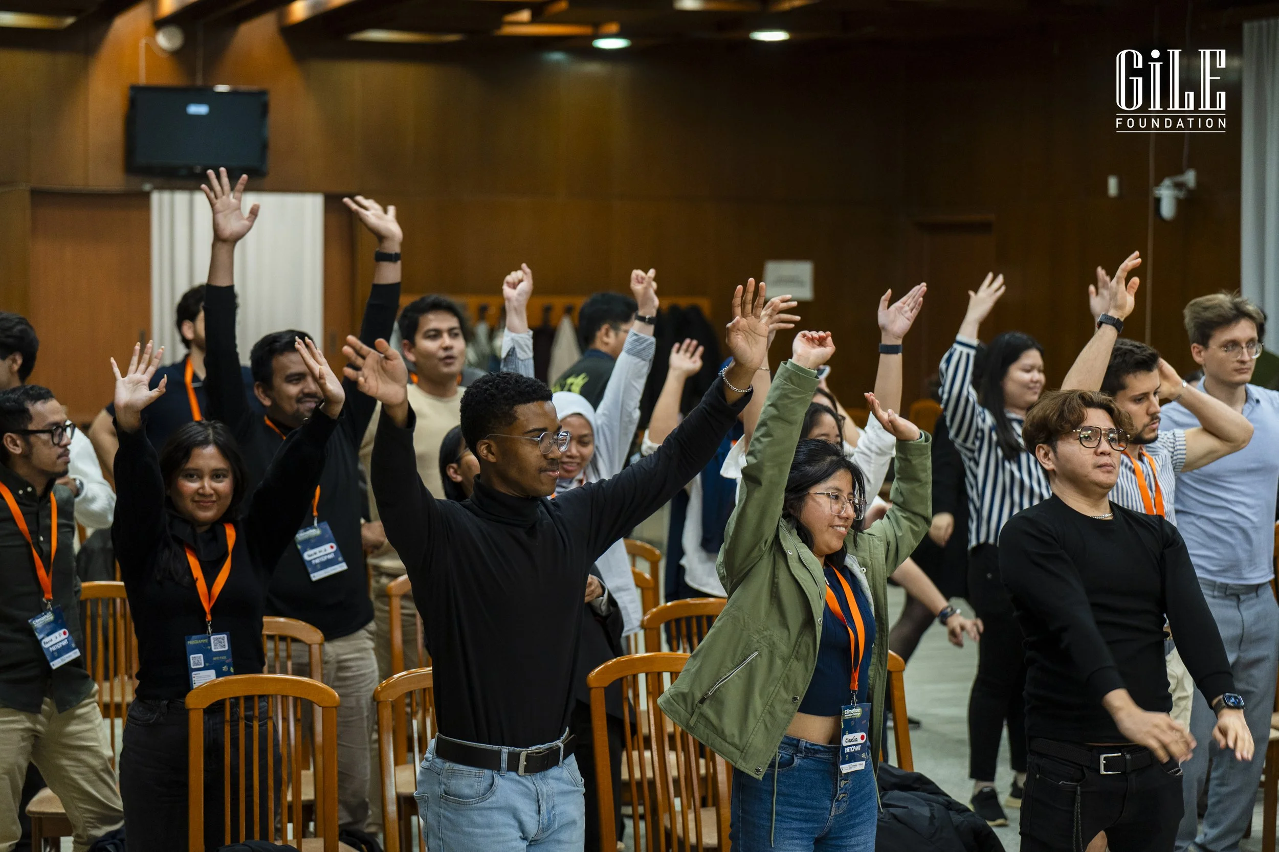 Group of young adults standing and raising their hands during an event in a wooden-finished room, with some wearing conference badges.