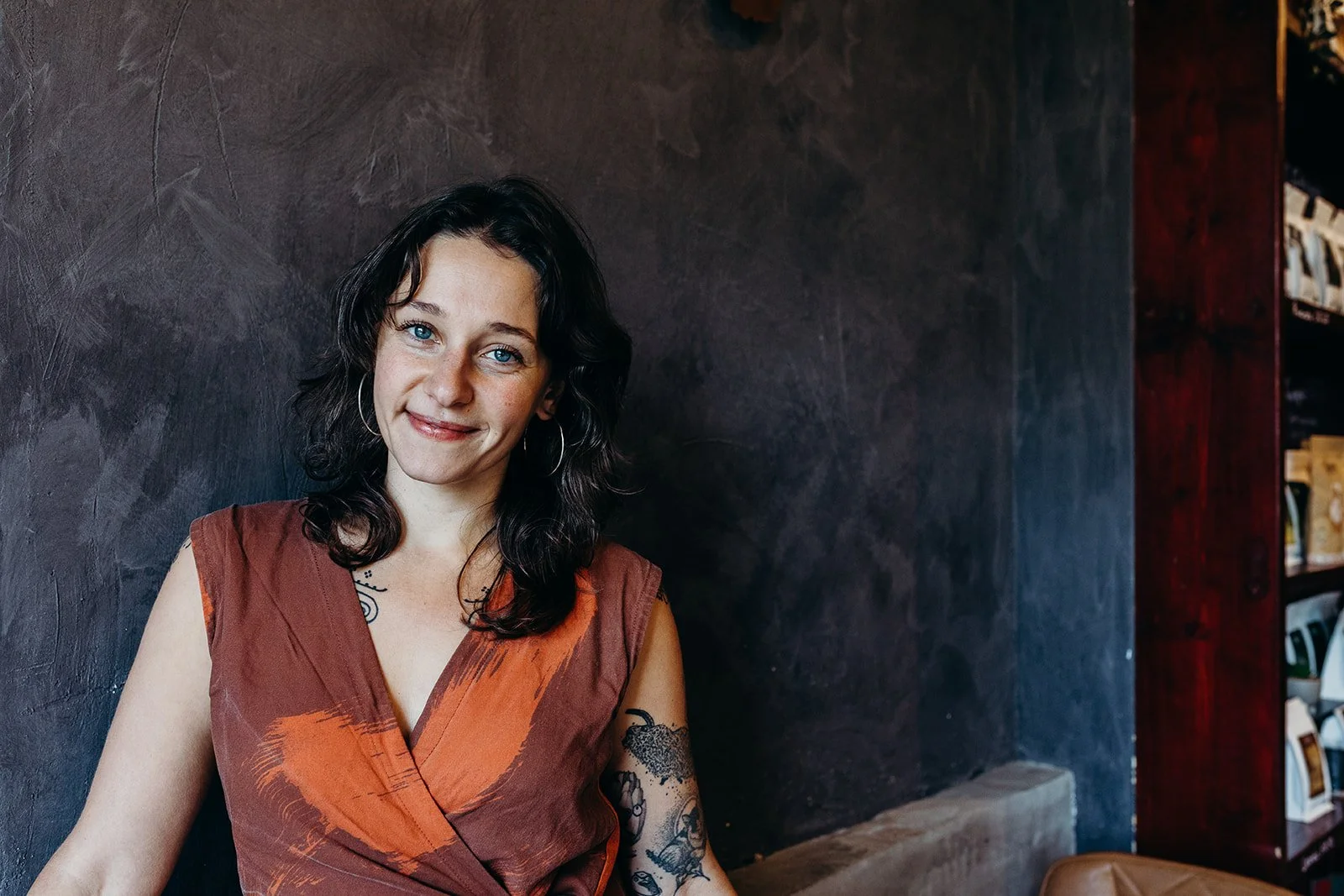 A woman with dark wavy hair, blue eyes, wearing hoop earrings and a sleeveless rust-colored top, standing indoors against a dark textured wall.