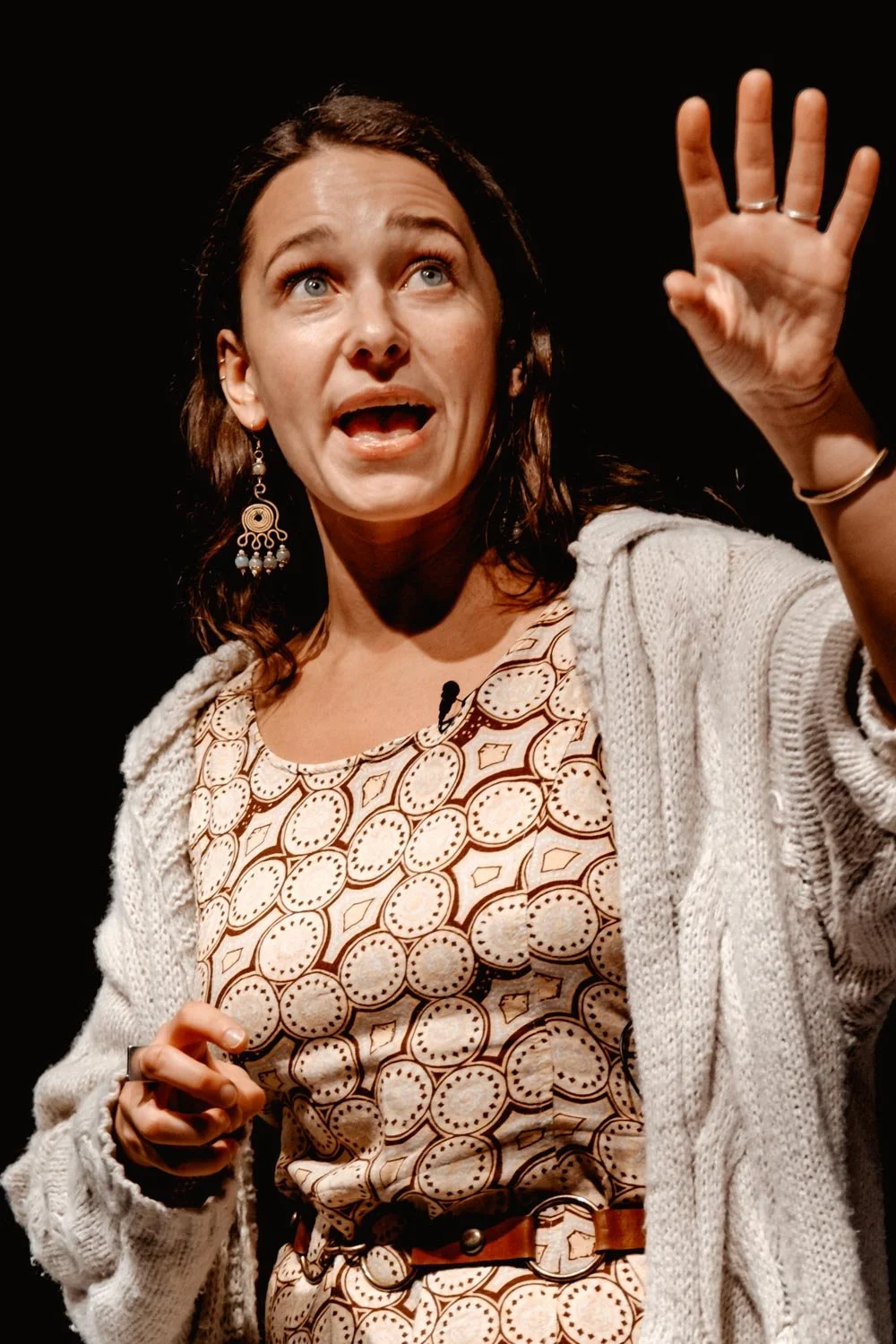 A woman with dark brown hair, wearing earrings and a patterned dress, appears to be speaking or explaining something while raising her right hand with fingers spread.