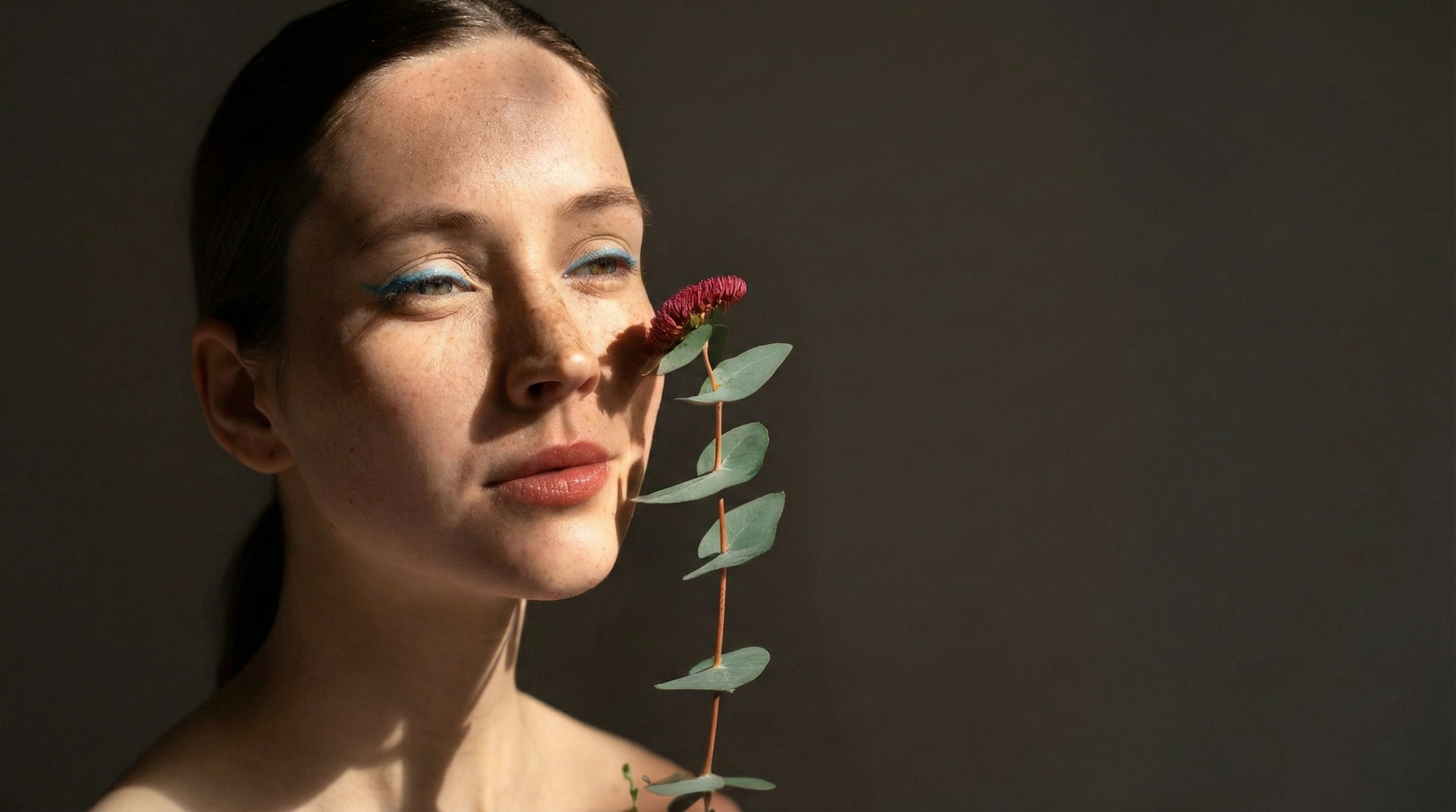 Ragazza con trucco evidenziato sugli occhi, che tiene una pianta con foglie e una spiga di fiori viola di fronte al viso, sfondo scuro.