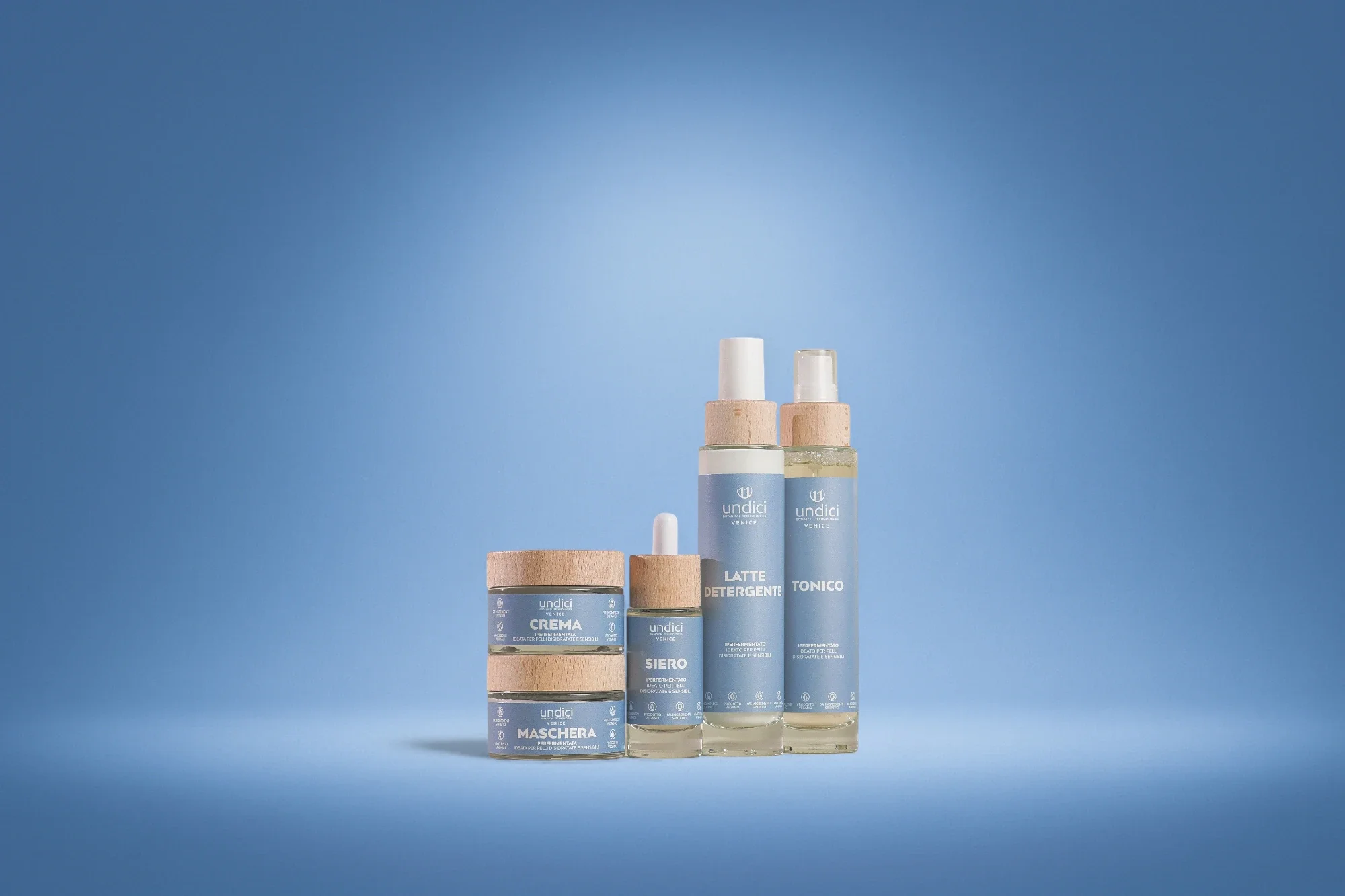 Set di prodotti per la cura della pelle con etichette blu e tappo di legno, tra cui crema, siero, latte detergente e tonico, disposti su sfondo blu