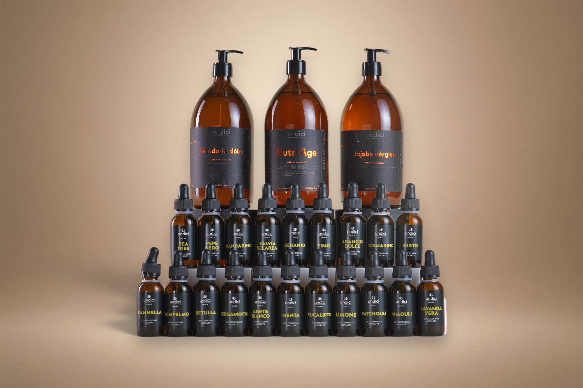 Set di bottiglie di oli essenziali e cosmetici naturali con etichette nere e testi in italiano, disposte in ordine, su uno sfondo beige.