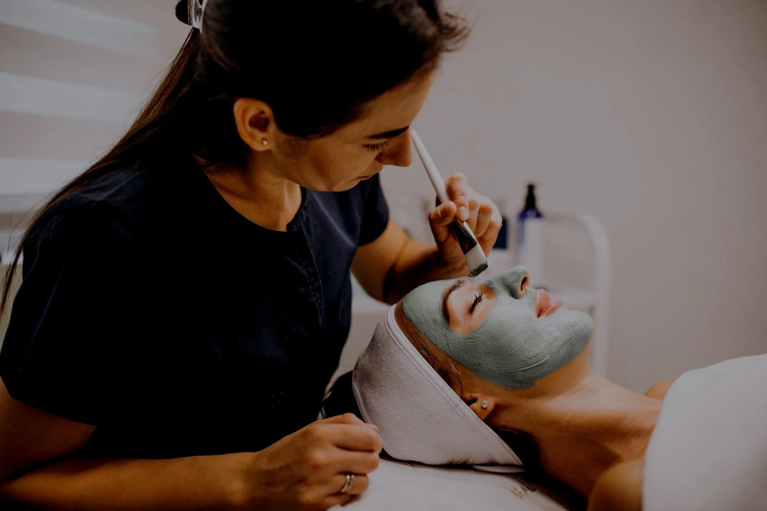 Una donna riceve un trattamento facciale con una maschera verde mentre giace con gli occhi chiusi, una professionista sta applicando il prodotto con un pennello in un centro estetico.