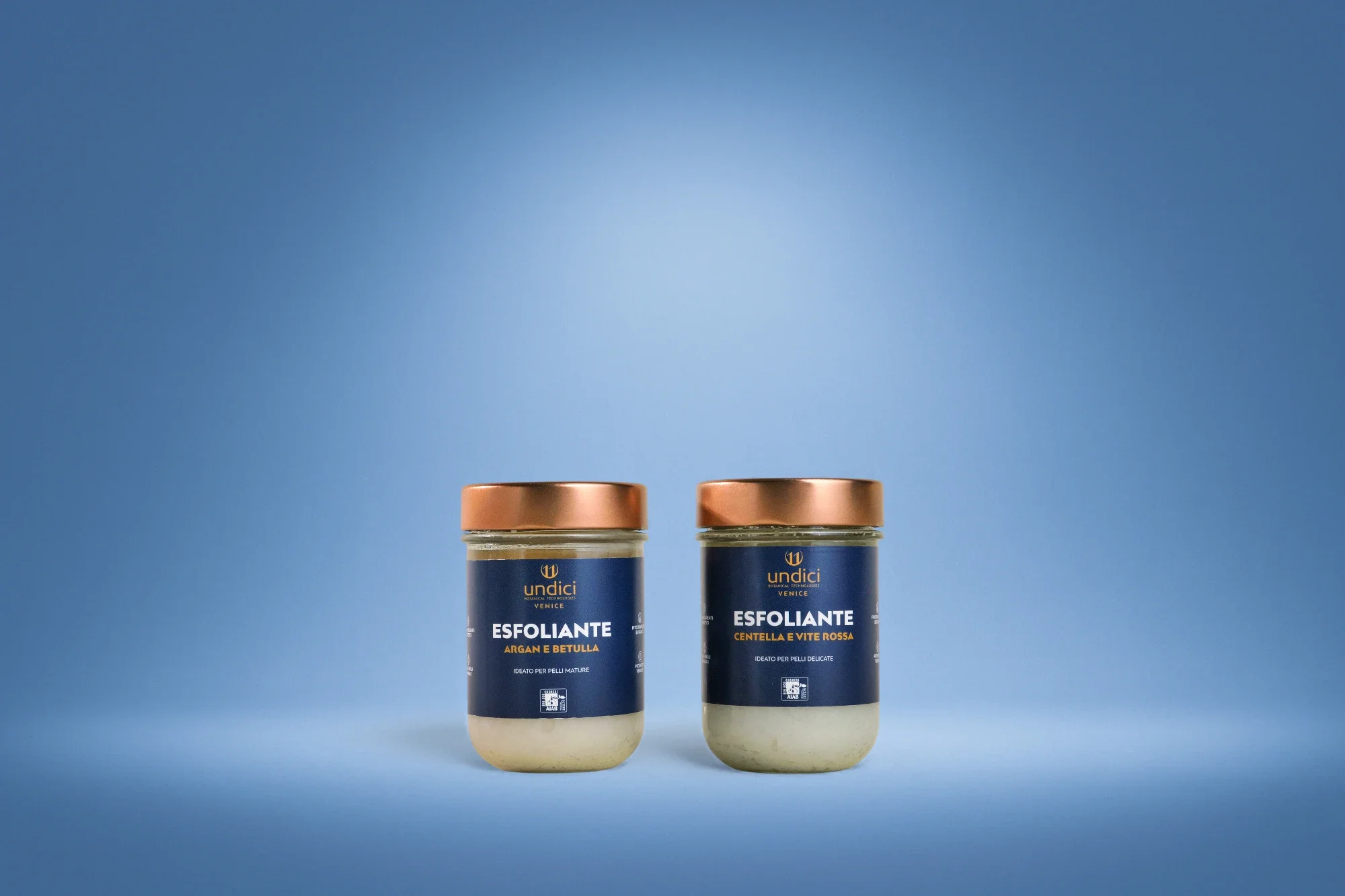 Due barattoli di scrub esfoliante di marca Undici, uno all'argan e betulla e l'altro alla centella e evita rossa, posizionati su uno sfondo blu uniforme