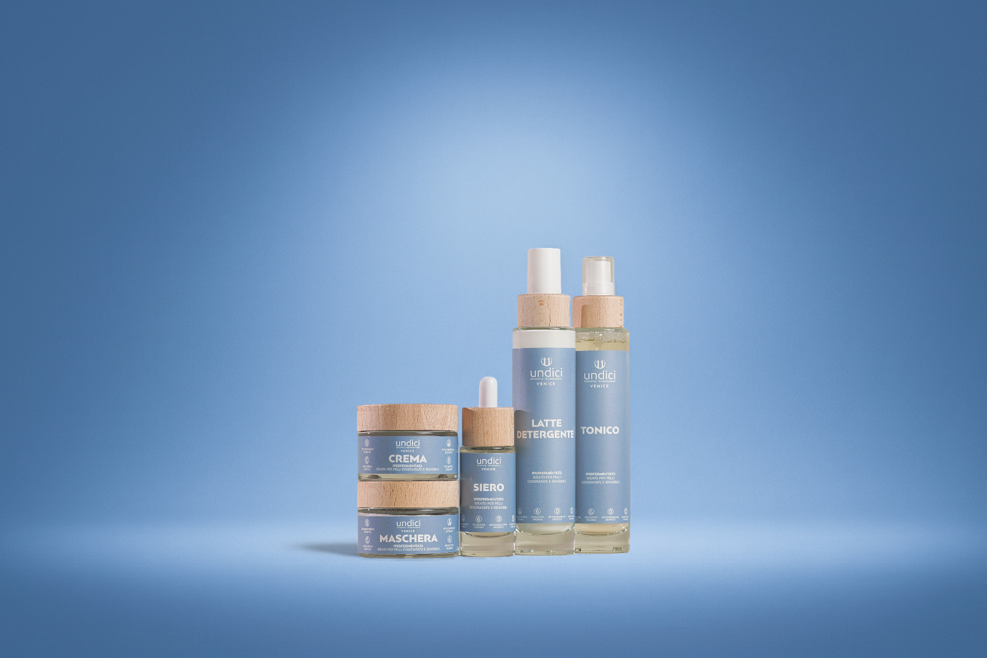 Set di cosmetici con confezioni blu e tappi di legno, tra cui crema, siero, latte detergente e tonico, su sfondo azzurro.