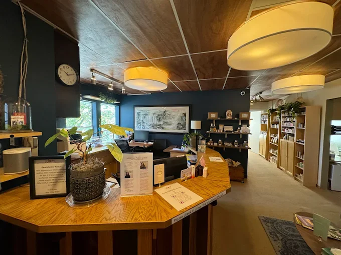 Interior of a cozy library or community center with a wooden reception desk, potted plant, large paintings, bookshelves, and warm lighting.