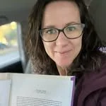 Woman with glasses holding a paper or document, smiling in a car.