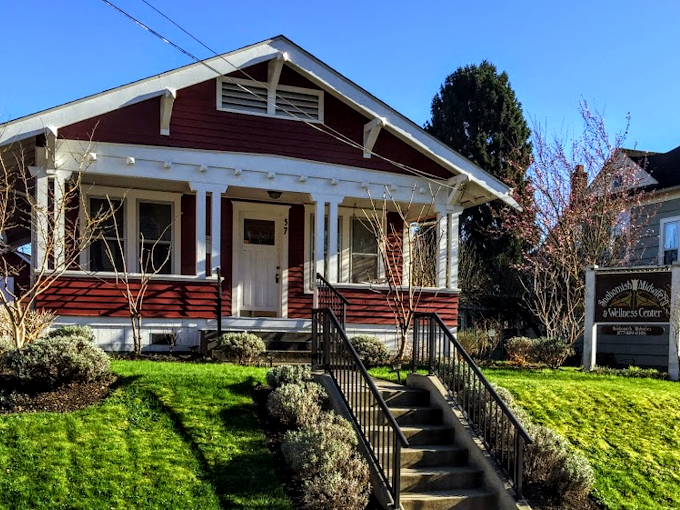 Red and white house with a front porch, stairs, and greenery, next to a wellness center sign in a sunny neighborhood.