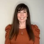 A woman with long brown hair smiling, wearing an orange top and a necklace, standing against a plain gray background.
