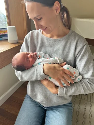 Woman holding a newborn baby in her arms, smiling and looking at the baby, inside a cozy room with a wooden window and a white speaker on the windowsill.