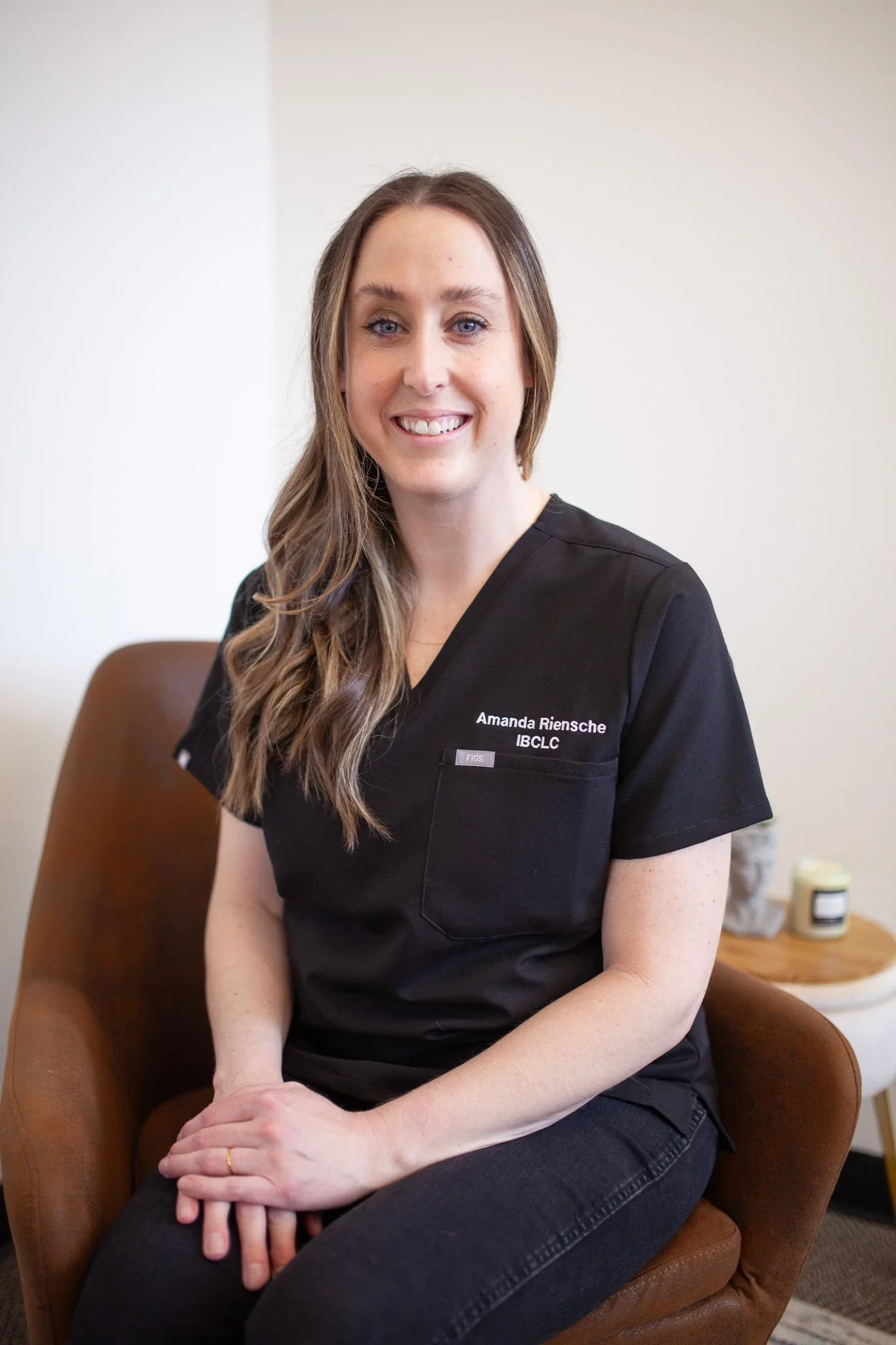 A woman sitting on a brown chair, wearing a black uniform with the name tag "Amanda Riensche" and the title "IBCLC". She has long, wavy brown hair and is smiling.