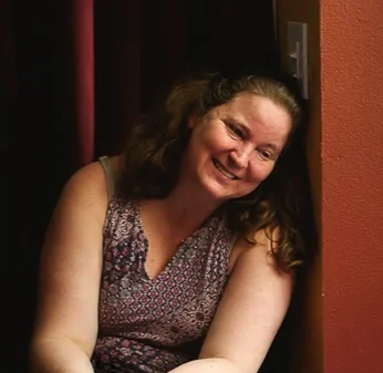 A smiling woman with curly brown hair sits against a brick wall.