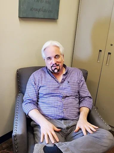 A man with white hair and a beard, wearing a purple checkered shirt, sitting in a gray armchair, in an office setting.