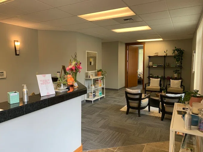 Reception area in a clinic or office with a counter, flower arrangements, seating area with chairs and a shelf with decorative plants and books.