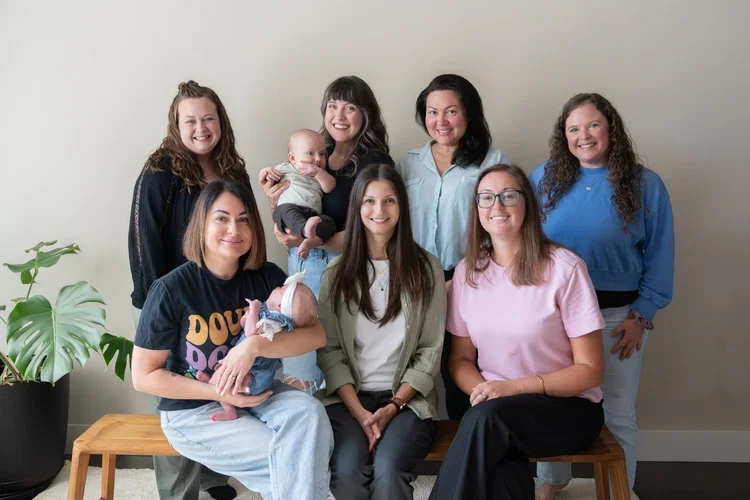 Group of seven young women, including one holding a baby and another holding a toddler, posing together in front of a plain wall with a plant nearby, smiling for the camera.