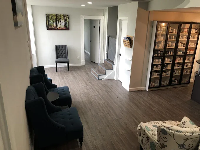 Empty room with four black chairs arranged along the wall and a patterned armchair in the corner. There is a painting of trees on the wall and a glass display case filled with pharmacy items. Stairs lead to the upper level, and a doorway leads to another room.