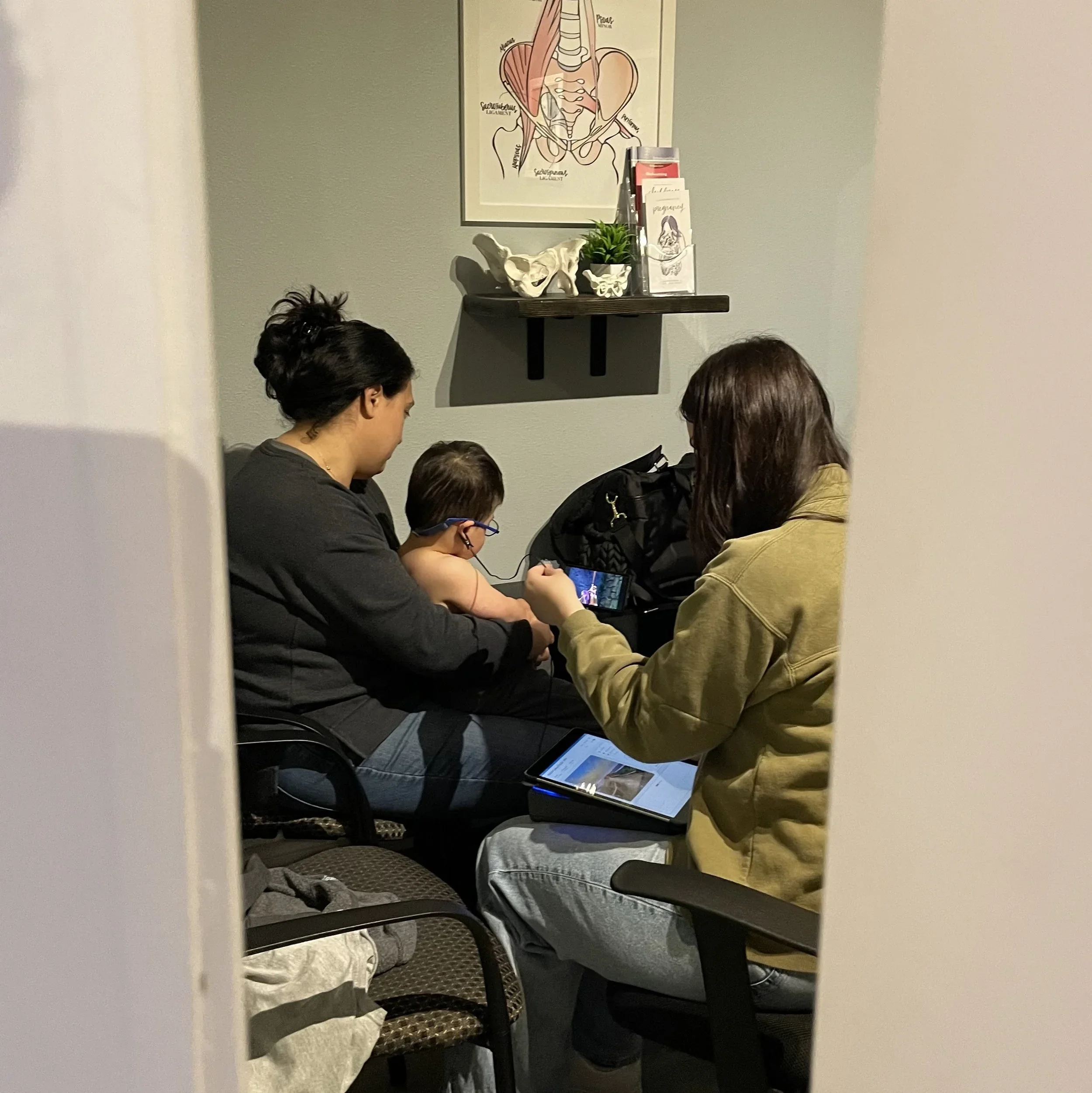 A woman and a child sitting in a waiting room, watching a tablet, with another woman showing something on her phone to the child. The room has a framed anatomy poster, a shelf with a model of a pelvis and some brochures.