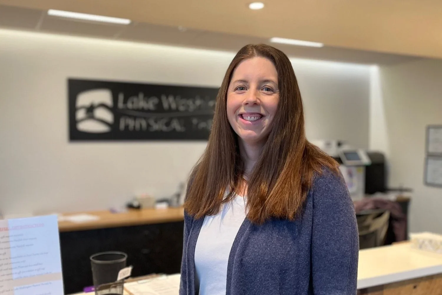 A smiling woman with long brown hair wearing a white shirt and a dark blazer standing in a reception area with a sign that says "Lake West Physical Therapy" in the background.