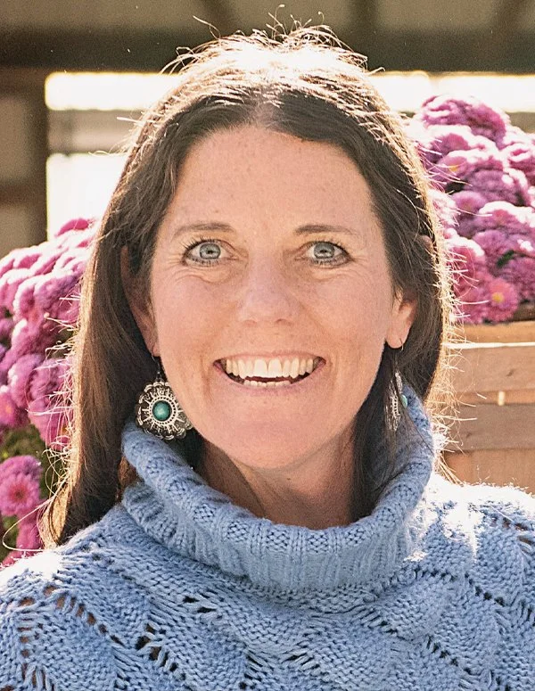 A woman with long brown hair and blue eyes smiling at the camera. She is wearing a light blue knitted turtleneck sweater and large, decorative earrings with turquoise stones. Behind her, there are pink flowers in a wooden crate, and sunlight is shining on her head, giving a bright outdoor setting.