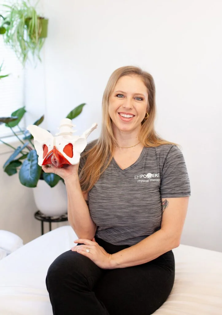 Woman smiling and holding a pelvis model in a clinical setting with plants in the background.