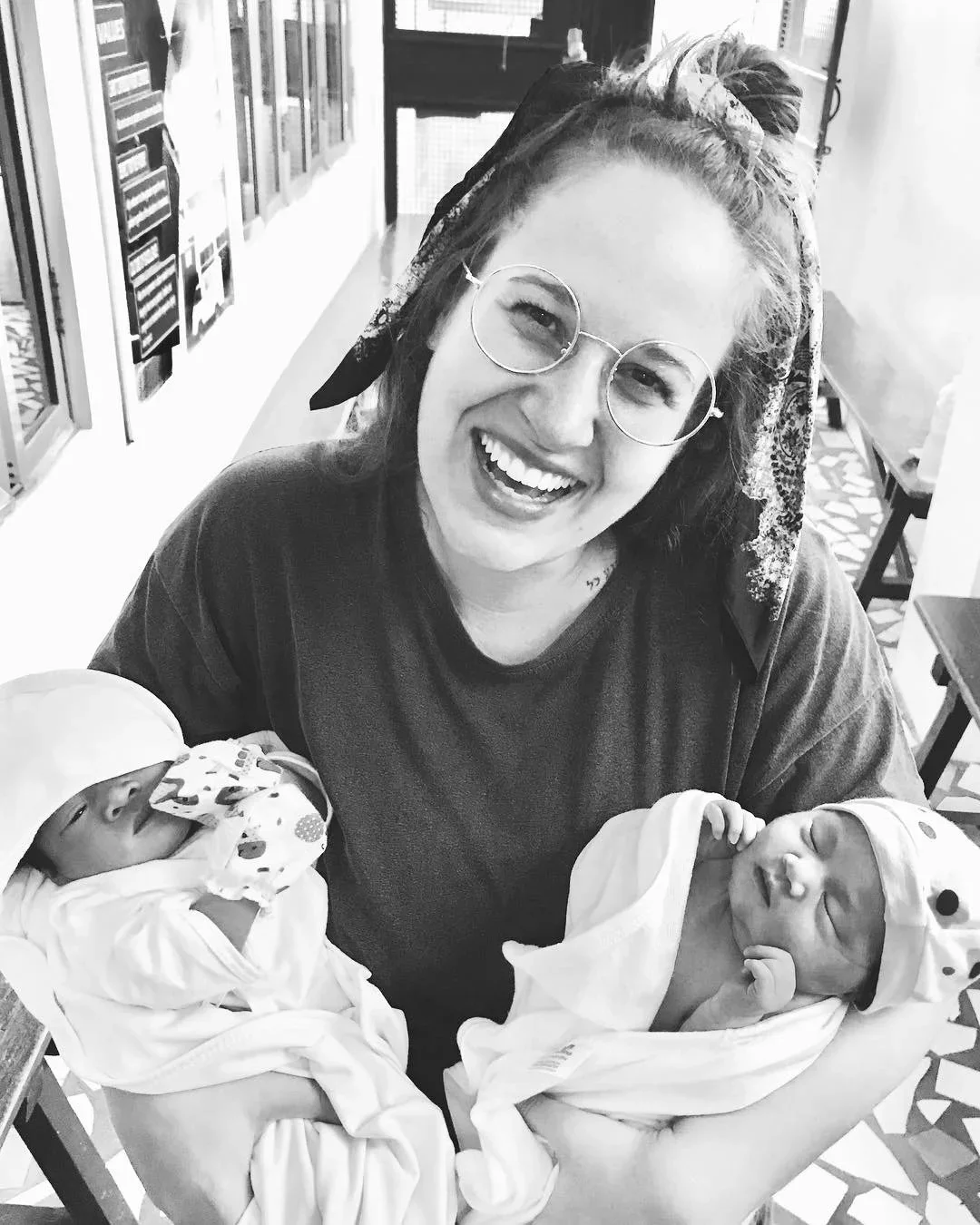 A woman with glasses and a patterned headscarf is smiling and holding two newborn babies, one in each arm, in a hospital room.