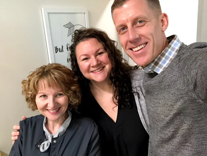 Three people smiling and taking a selfie indoors, with a framed picture on the wall in the background.