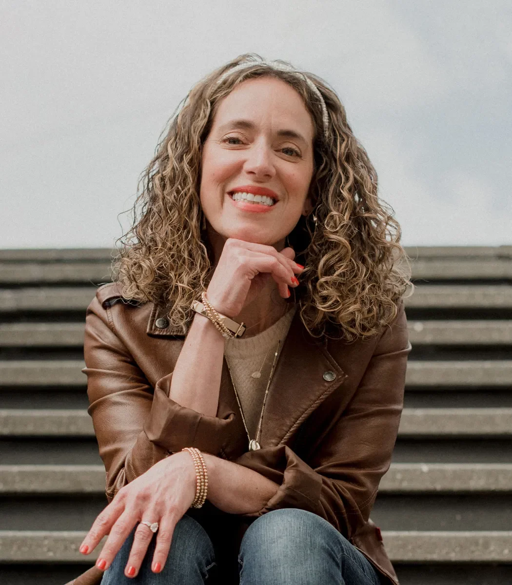 A woman with curly hair sitting on steps, smiling and resting her chin on her hand, wearing a brown leather jacket and jewelry.