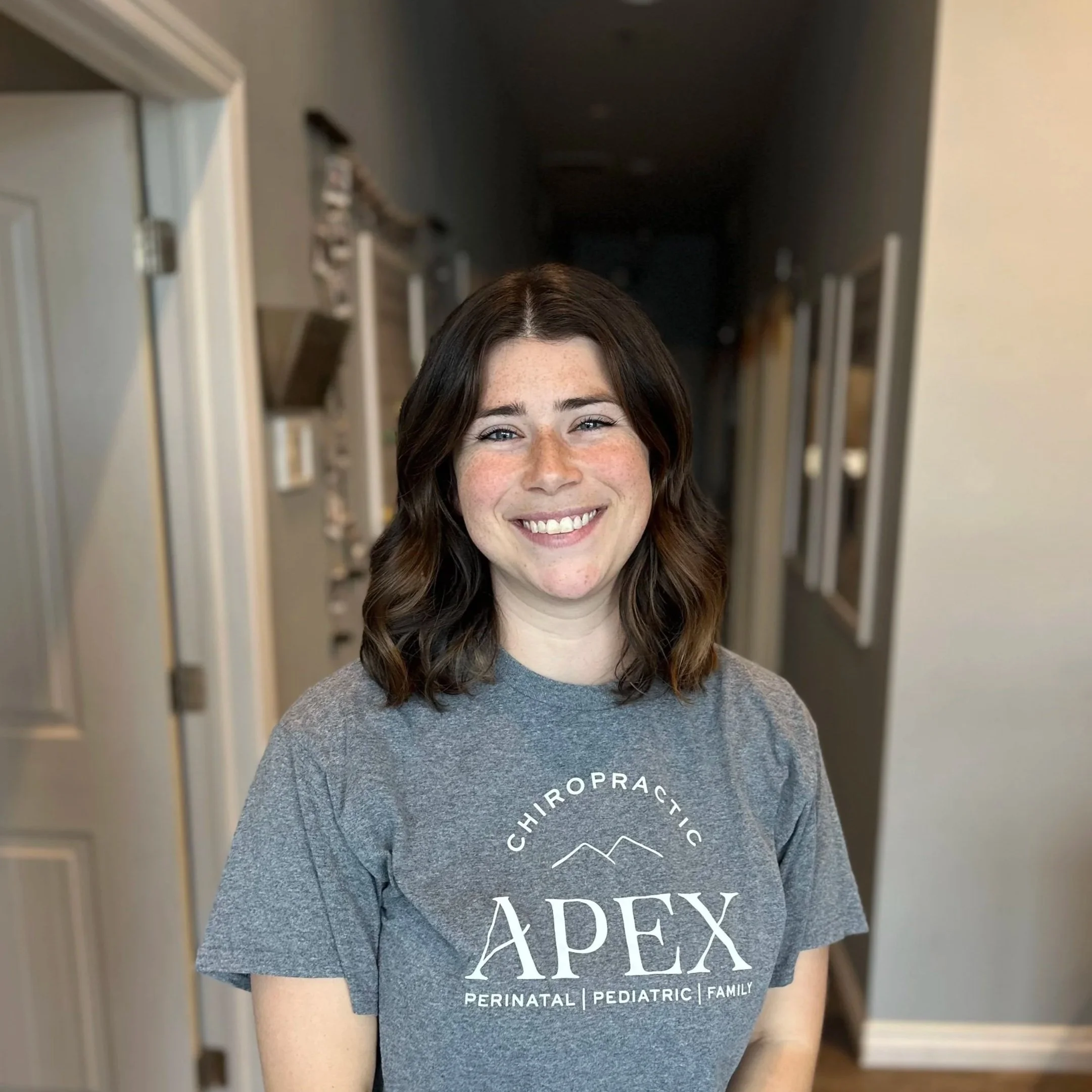 A young woman with shoulder-length wavy brown hair, smiling, wearing a gray t-shirt that says 'Chiropractic APEX Perinatal Pediatric Family'.