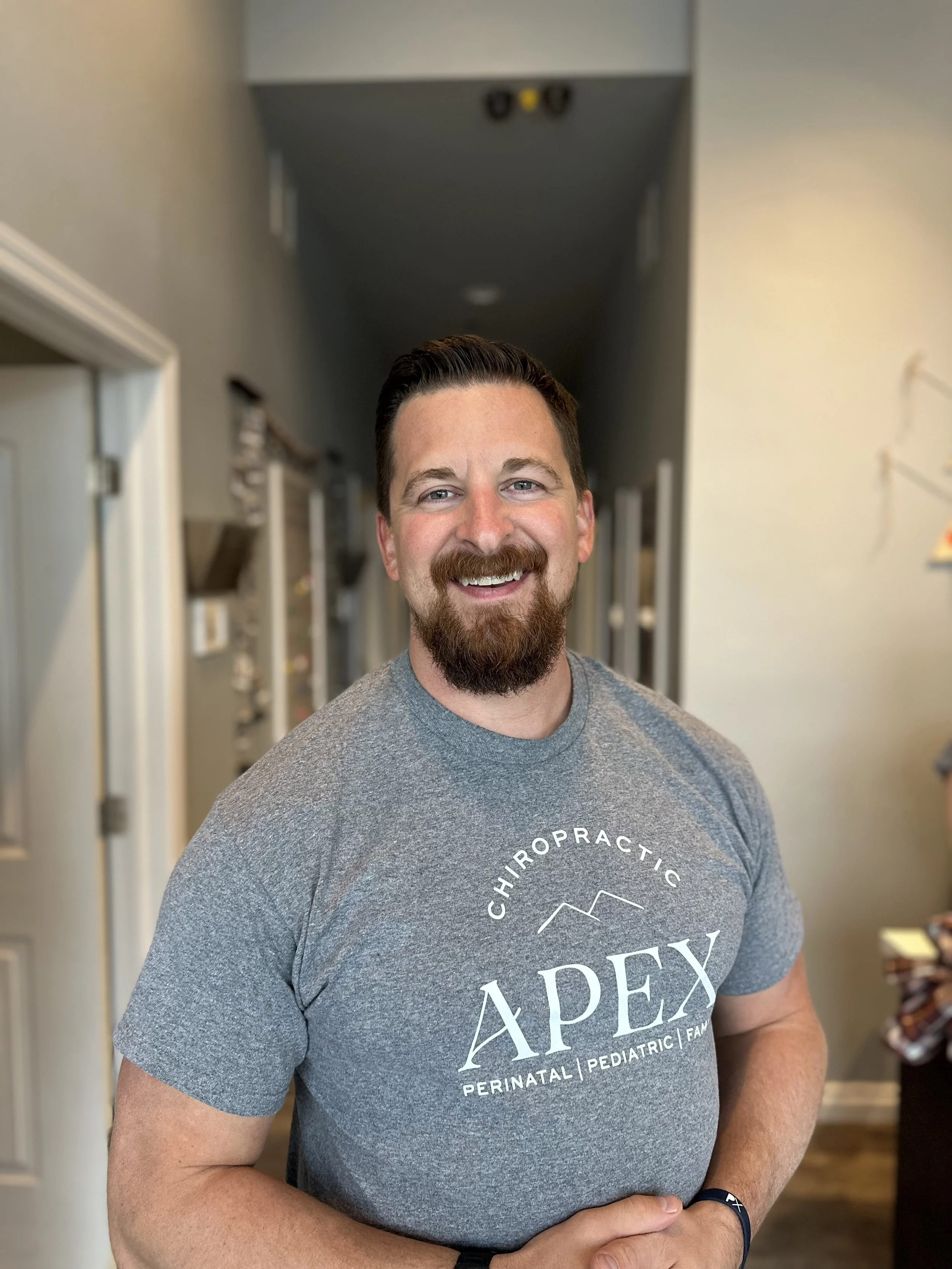 A smiling man with a beard and short dark hair standing indoors in front of a blurred background. He is wearing a gray T-shirt with the words "Chiropractic Apex Perinatal Pediatric Family" printed on it.