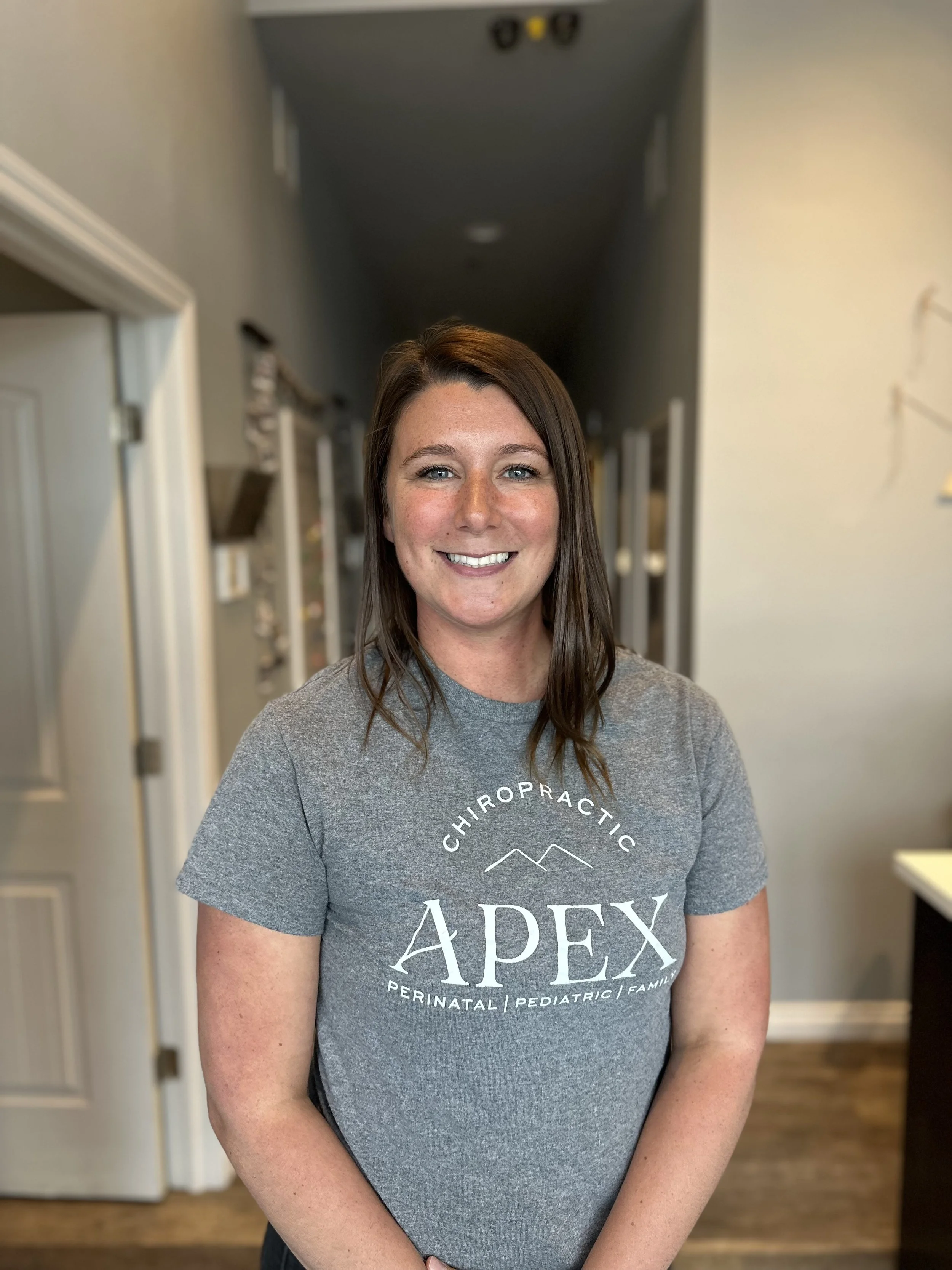 A woman with brown hair and blue eyes smiling at the camera inside a room, wearing a gray t-shirt with the logo 'APEX Chiropractic Perinatal Pediatric Family' on it.