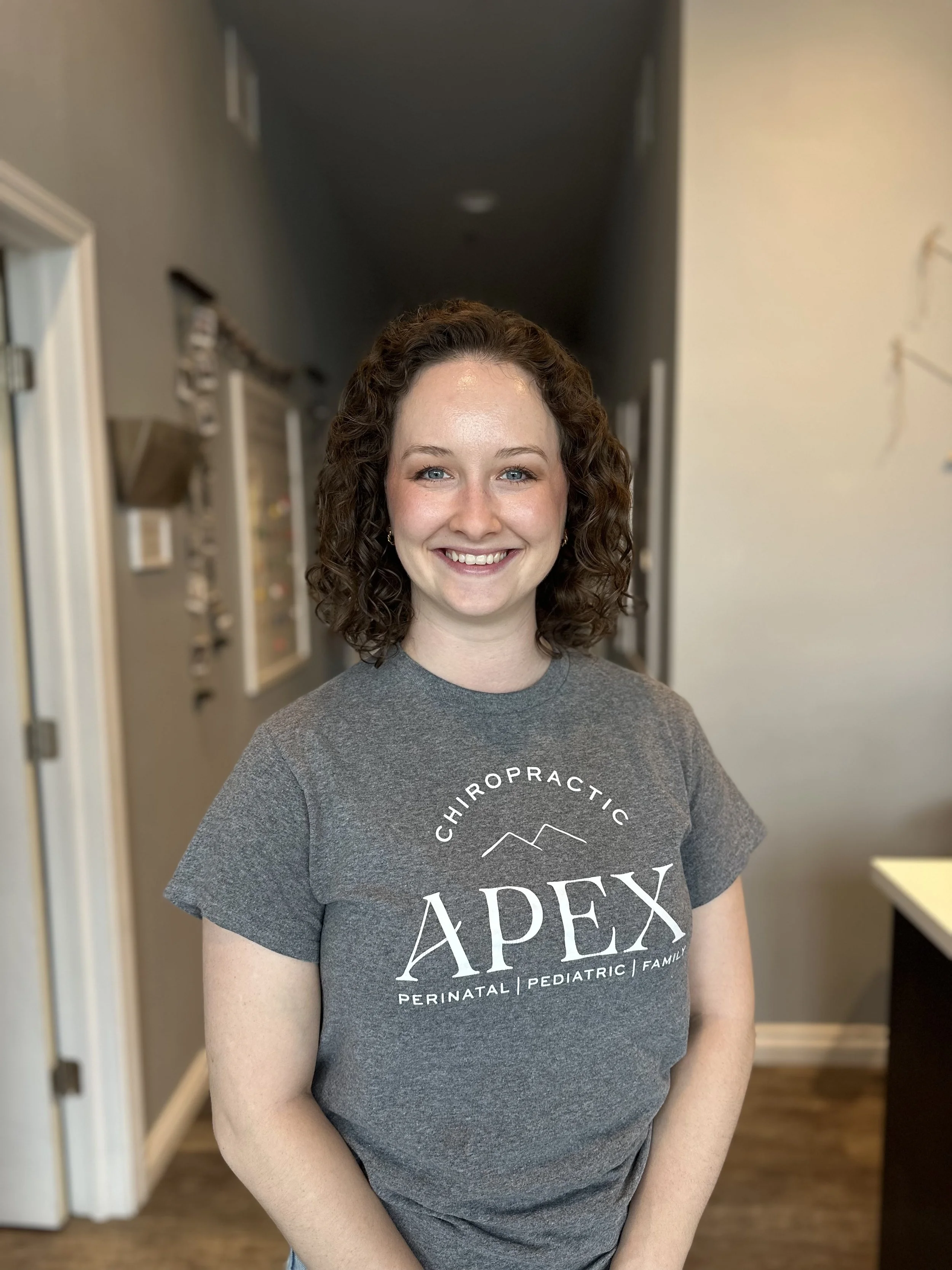 A woman with curly brown hair and a gray t-shirt that says  APPEX Chiropractic, Perinatal, Pediatric, Family, smiling in an indoor setting.