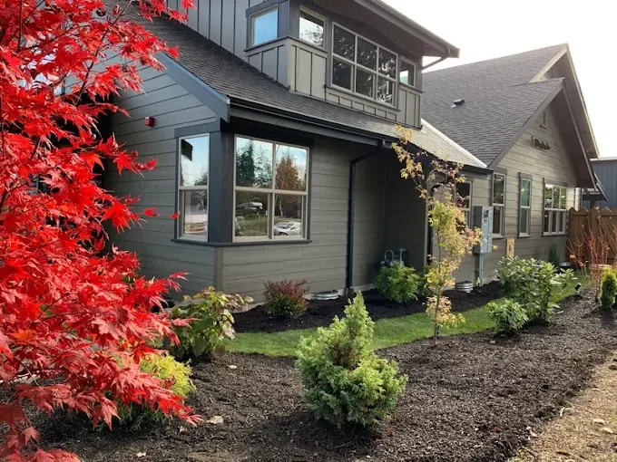 A modern gray house with multiple windows, surrounded by a landscaped garden with various shrubs and trees, including a red-leafed tree in the foreground.