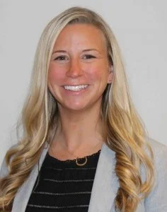 A smiling woman with long blonde hair, wearing a black top and a gray blazer, against a plain background.