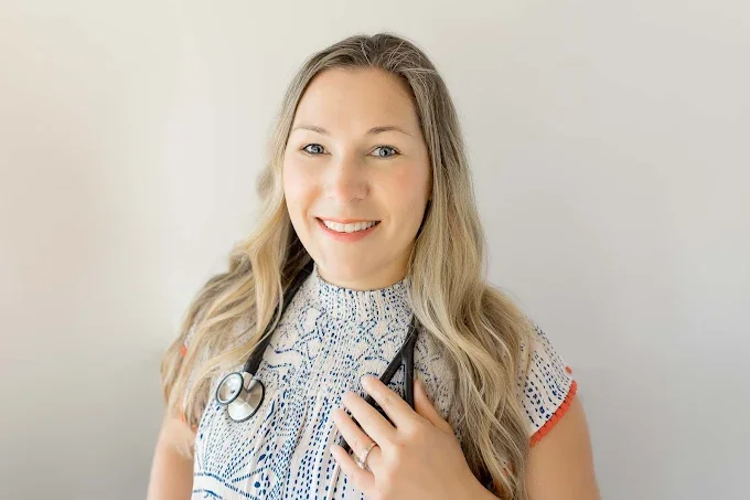 Smiling female doctor with blonde hair, wearing a patterned blouse and stethoscope around her neck, standing against a plain white background.
