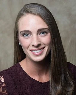 Portrait of a young woman with long brown hair, smiling, wearing a dark purple top, and standing against a neutral background.