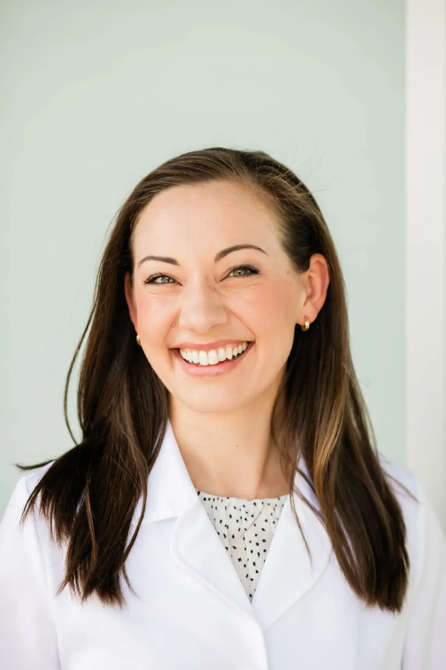 A smiling woman with brown hair, wearing a white lab coat and small hoop earrings, standing against a light-colored background.