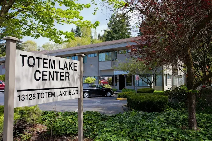 Sign for Totem Lake Center with address 13128 Totem Lake Blvd in front of a two-story commercial building surrounded by greenery and parked cars.