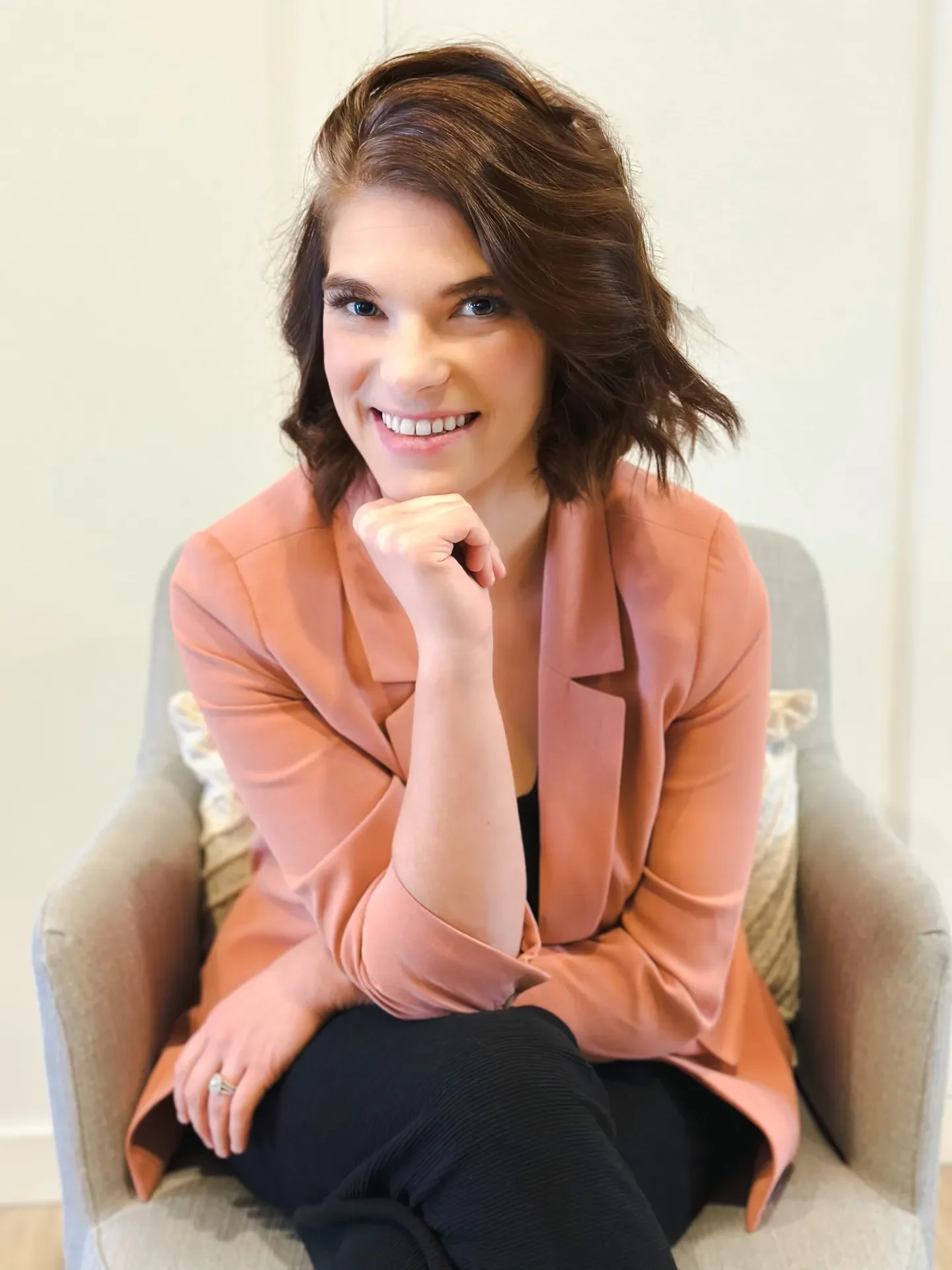 A smiling woman with shoulder-length brown hair, wearing a peach-colored blazer, sitting on a beige armchair with a patterned pillow, in a well-lit room.