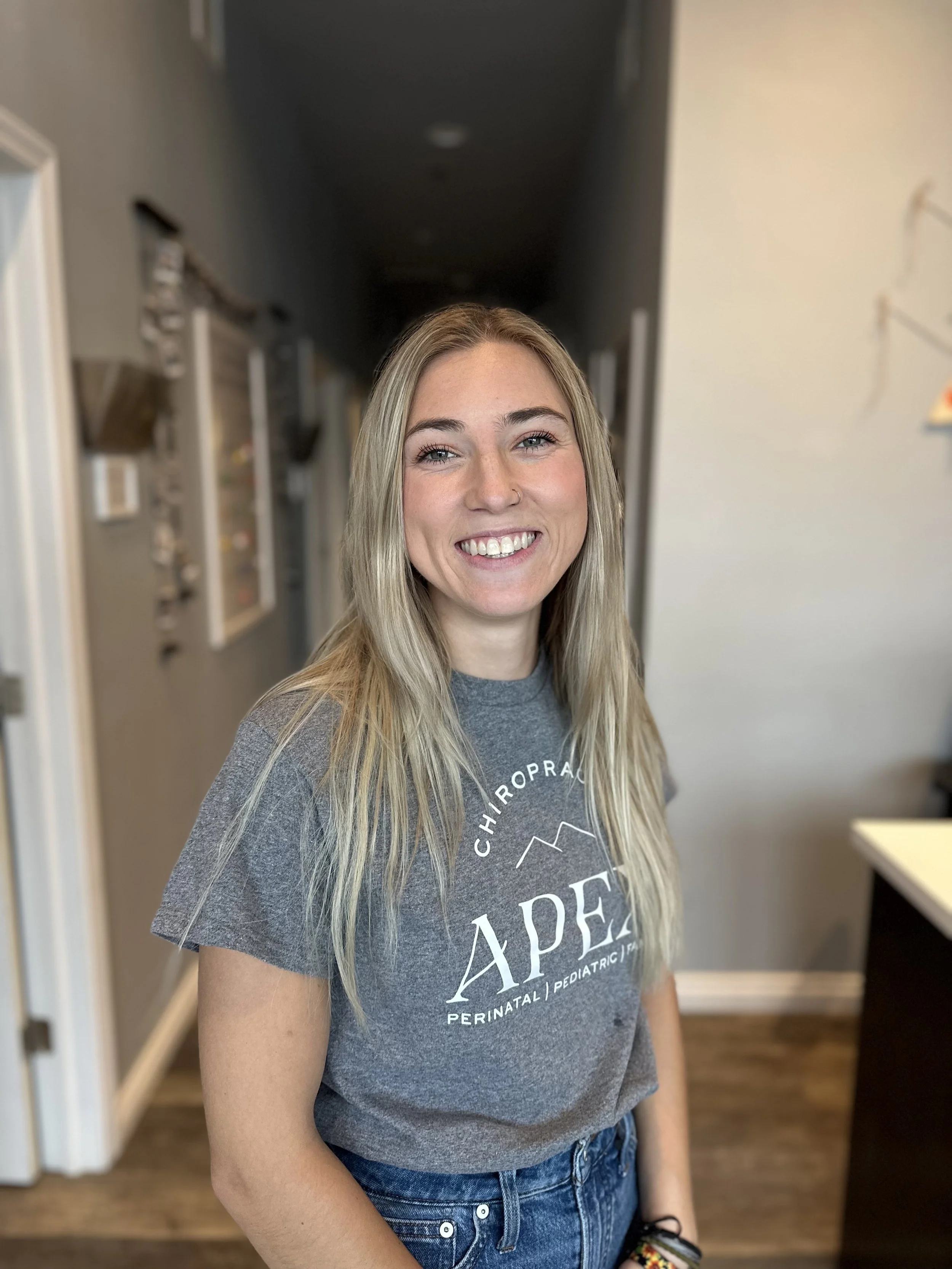 Smiling young woman with long blonde hair wearing a gray T-shirt and blue jeans inside a home.