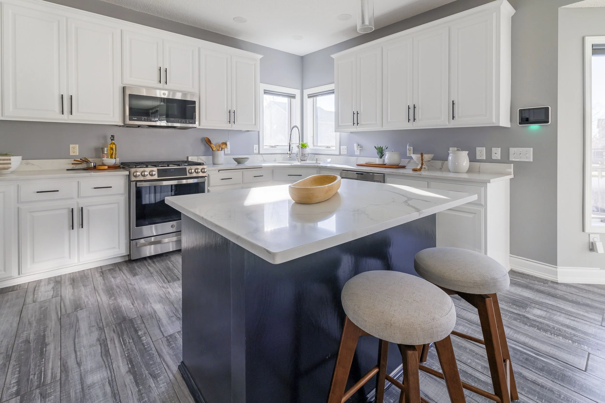 Modern kitchen with white cabinets, stainless steel appliances, a central island with a dark base and white countertop, gray walls, and gray wood-look flooring. There are two bar stools with beige cushions in front of the island.