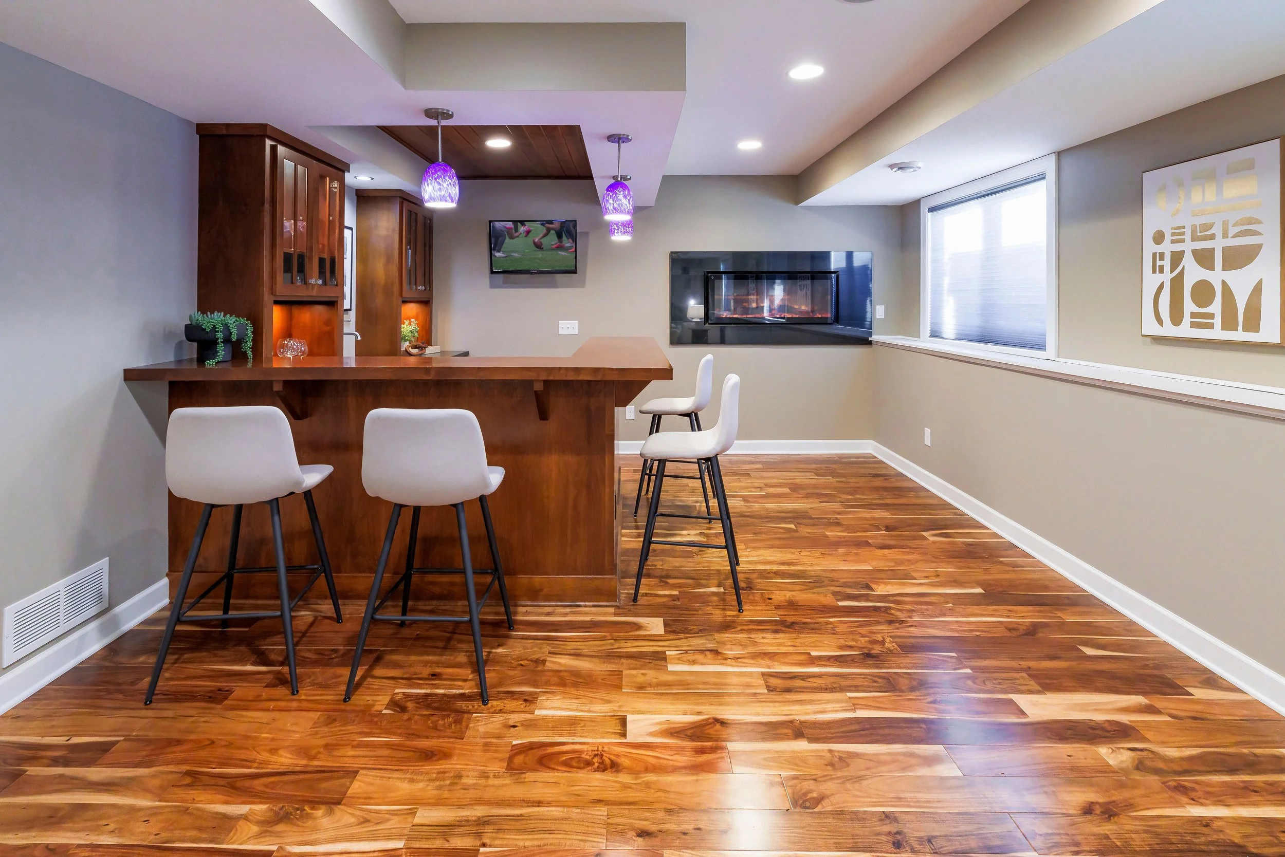 Interior view of a modern living space with a wooden flooring, a bar counter with white chairs, and a wall-mounted television. There is a fireplace on the wall, a large window with blinds, and a piece of abstract art on the wall.